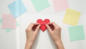 A bright, minimalist flat-lay of hands folding a red origami heart from a square sheet of paper on a clean white desk, with a few colorful paper squares around it in soft natural light.