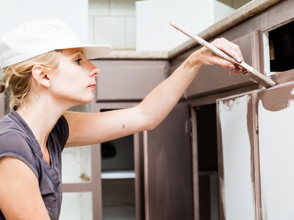 An image of a woman painting a cabinet for an article on how to paint kitchen cabinets.